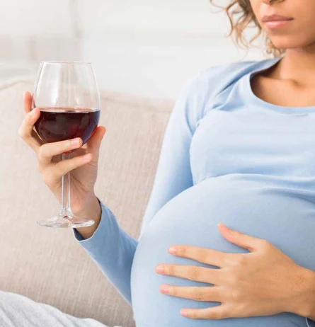 Schwangere Person in hellblauem Kleid hält ein Glas Rotwein und streichelt ihren Bauch.