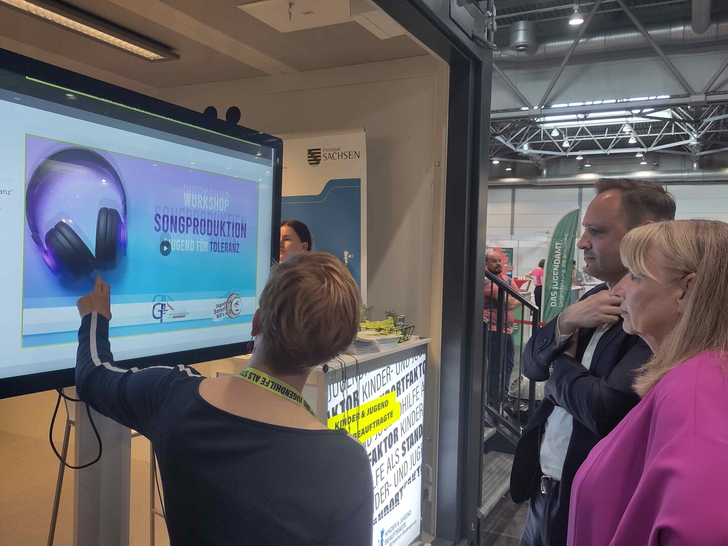 Auf diesem Foto sieht man den Messestand der Kinder- und Jugendbeauftragten der Säsischen Staatsregierung Susann Rüthrich. Davor steht sie zusammen mit Petra Köpping, der Sozialministerin und dem Landtagspräsidenten Alexander Dirks. Sie schauen sich auf d