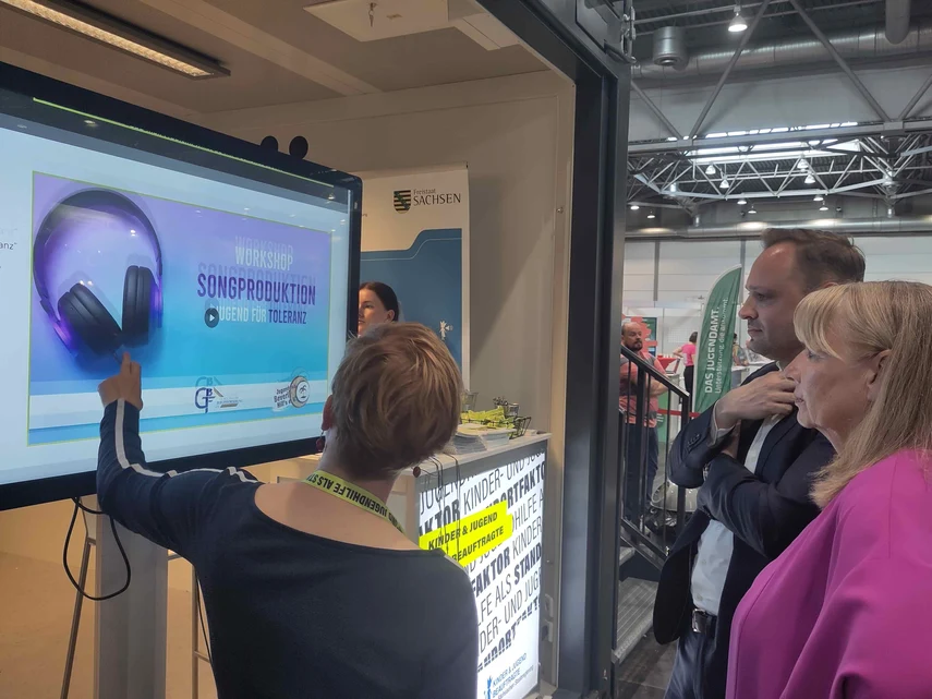 Auf diesem Foto sieht man den Messestand der Kinder- und Jugendbeauftragten der Säsischen Staatsregierung Susann Rüthrich. Davor steht sie zusammen mit Petra Köpping, der Sozialministerin und dem Landtagspräsidenten Alexander Dirks. Sie schauen sich auf d
