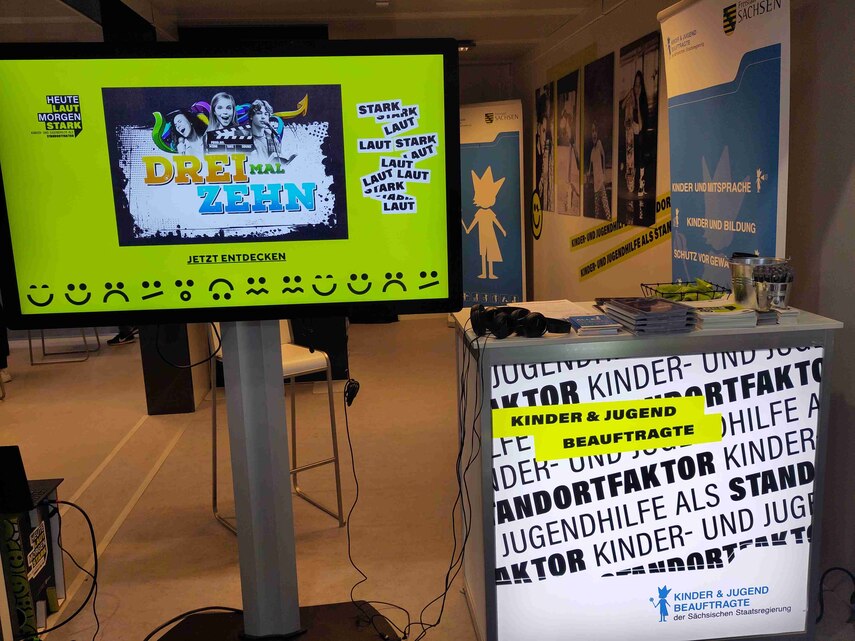 Auf diesem Foto ist der Messestand der Kinder- und Jugendbeauftragten zu sehen. Auf einem großen Bildschirm sieht man Beiträge des Wettbewerbs 3mal10. Auf einem Tisch, rechts daneben, sind Werbematerialien und die Texte des Wettbewerbs ausgelegt.