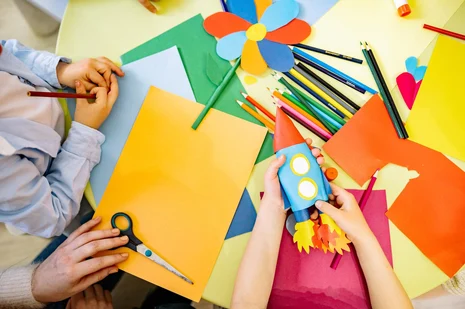 Zwei Kinder basteln an einem Tisch. Auf dem Tisch liegen viele Buntstifte und buntes Bastelpapier. Das eine Kind hält eine gebastelte Rakete in der Hand.