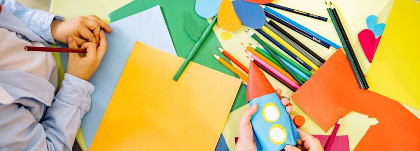 Draufsicht auf einen bunten Basteltisch, an dem ein Kind eine kleine Papierrakete in den Händen hält. Umgeben von farbigen Papierbögen, Buntstiften und einer gebastelten Papierblume.