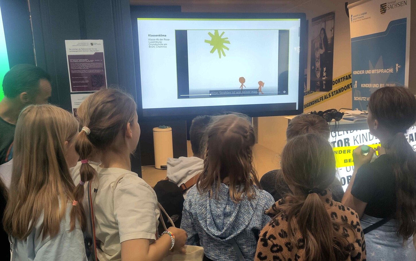 Auf diesem Bild ist eine Gruppe junger Kinder zu sehen, die auf einen Monitor schauen. Sie sind Teilnehmer des Wettbewerbs 3mal10 und schauen sich ihren Film auf dem Monitor an. Auf dem Monitor sieht man eine Sonne und zwei Kinder aus Papier. Der Titel de