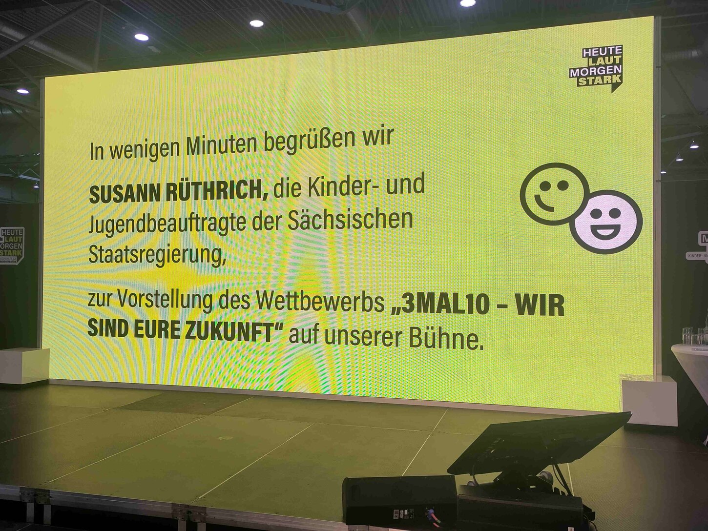 Auf diesem Foto sieht man die Sachsenbühne. Auf gelben Hintergrund steht in schwarzer Schrift geschrieben: "In wenigen Minuten begrüßen wir Susann Rüthrich, die Kinder- und Jugendbeauftragte der Sächsischen Staatsregierung, zur Vorstellung des Wettbewerbs