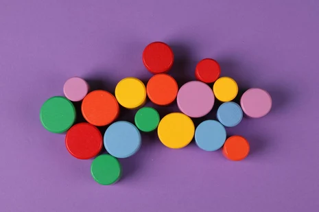 Draufsicht auf eine Gruppe verschieden großer, bunter Holzscheiben auf einem violetten Hintergrund. Die runden Spielsteine in den Farben Rot, Orange, Gelb, Grün, Hellblau und Rosa sind lose und leicht überlappend in der Bildmitte gruppiert.