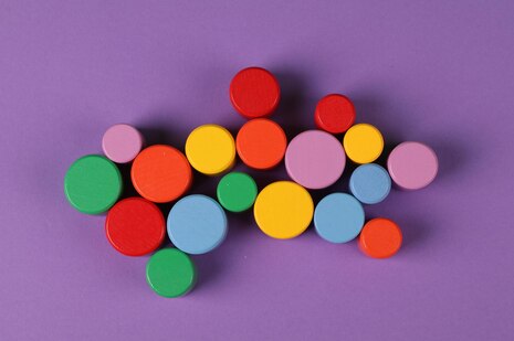 Draufsicht auf eine Gruppe verschieden großer, bunter Holzscheiben auf einem violetten Hintergrund. Die runden Spielsteine in den Farben Rot, Orange, Gelb, Grün, Hellblau und Rosa sind lose und leicht überlappend in der Bildmitte gruppiert.