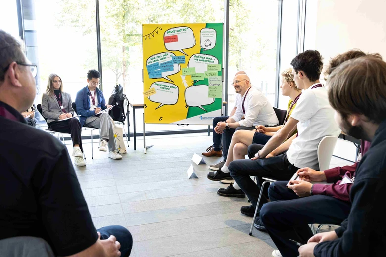 junge Menschen sitzen im Kreis und diskutieren mit Staatssekretär Kralinski. Im Hintergrund eine Pinnwand mit Sprechblasen
