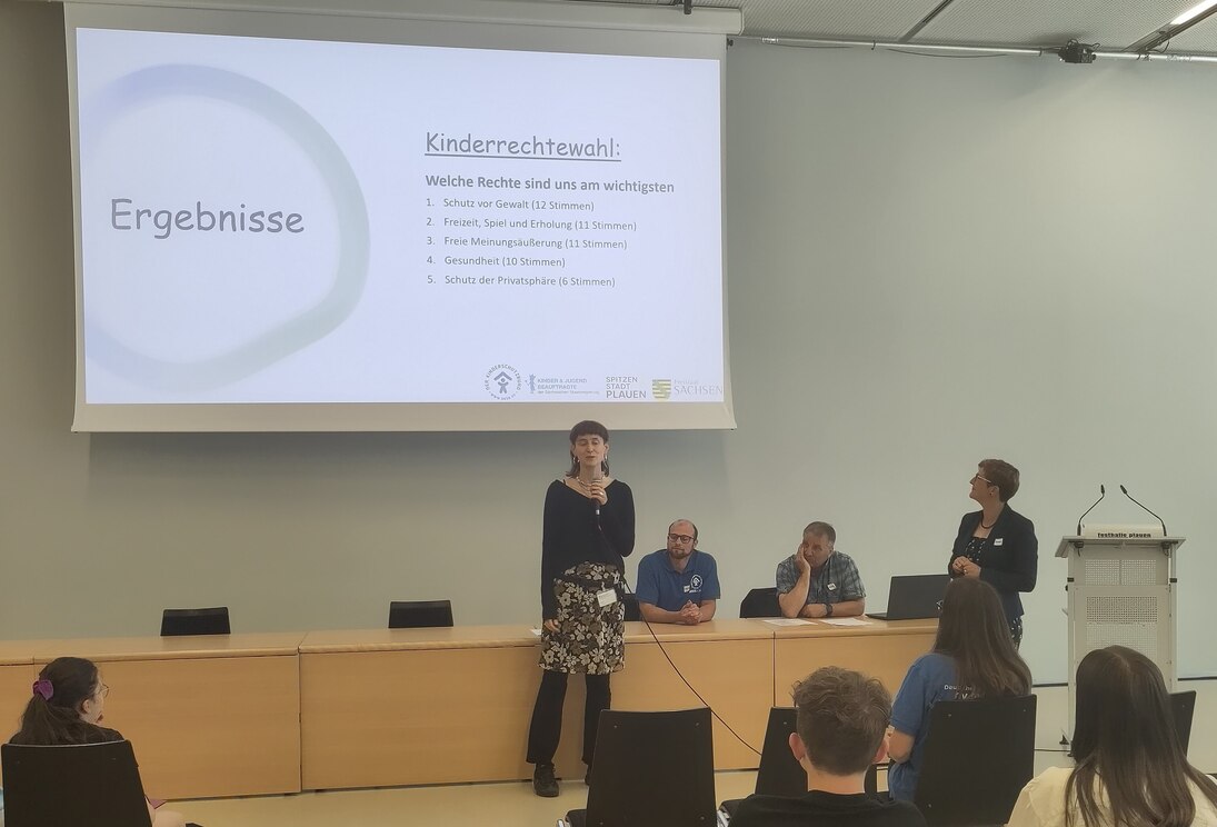 Person spricht vor Publikum. Im Hintergrund eine Leinwand mit dem Wort Kinderrechtewahl