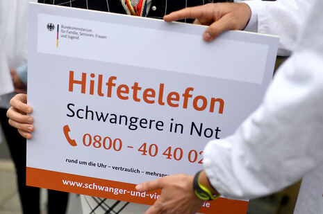 Ein Schild mit der Aufschrift "Hilfetelefon Schwangere in Not Telefon 0800 - 40 40 02.." wird von drei Händen gehalten
