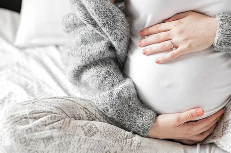 Junge Frau sitzt auf Bett, hält sich den schwangeren Bauch