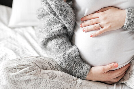 Junge Frau sitzt auf Bett, hält sich den schwangeren Bauch