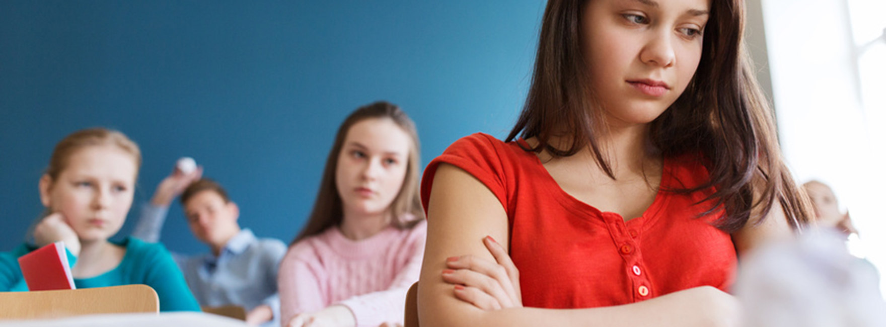 Eine Schülerin (Teenager) sitzt mit bedrücktem Gesichtsausdruck auf der Schulbank, hinter hier zwei Klassenkameradinnen, die besorgt zu ihr blicken