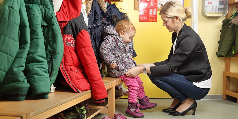 Ein Kind sitz in der Kita auf der Bank und wird von seiner Mutter angezogen.