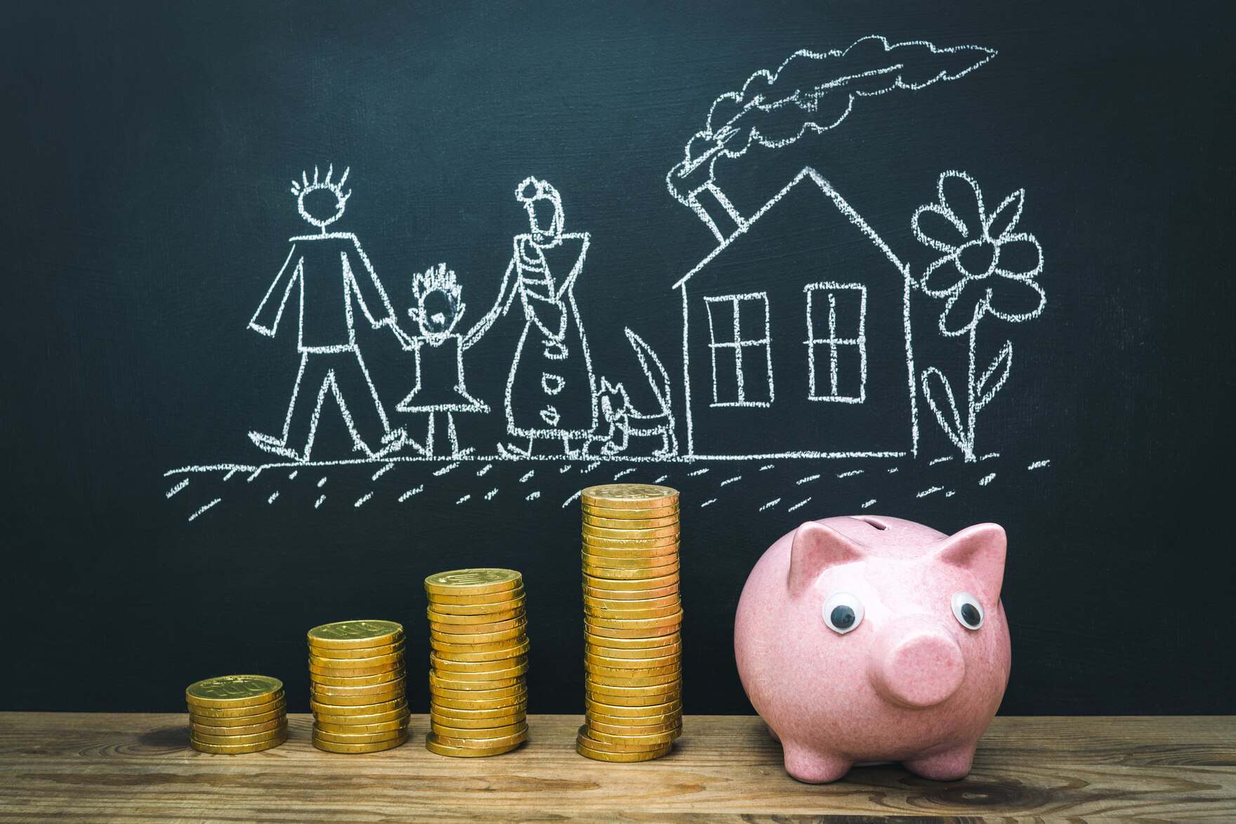 gestapelte Geldstücke neben einem rosa Sparschwein vor einer Kreistafel mit der Zeichnung einer Familie mit Haus