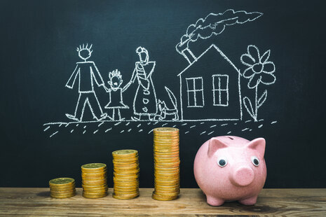 gestapelte Münzen neben einem rosa Sparschwein vor einer Kreidetafel mit einer gezeichneten Familie mit Haus