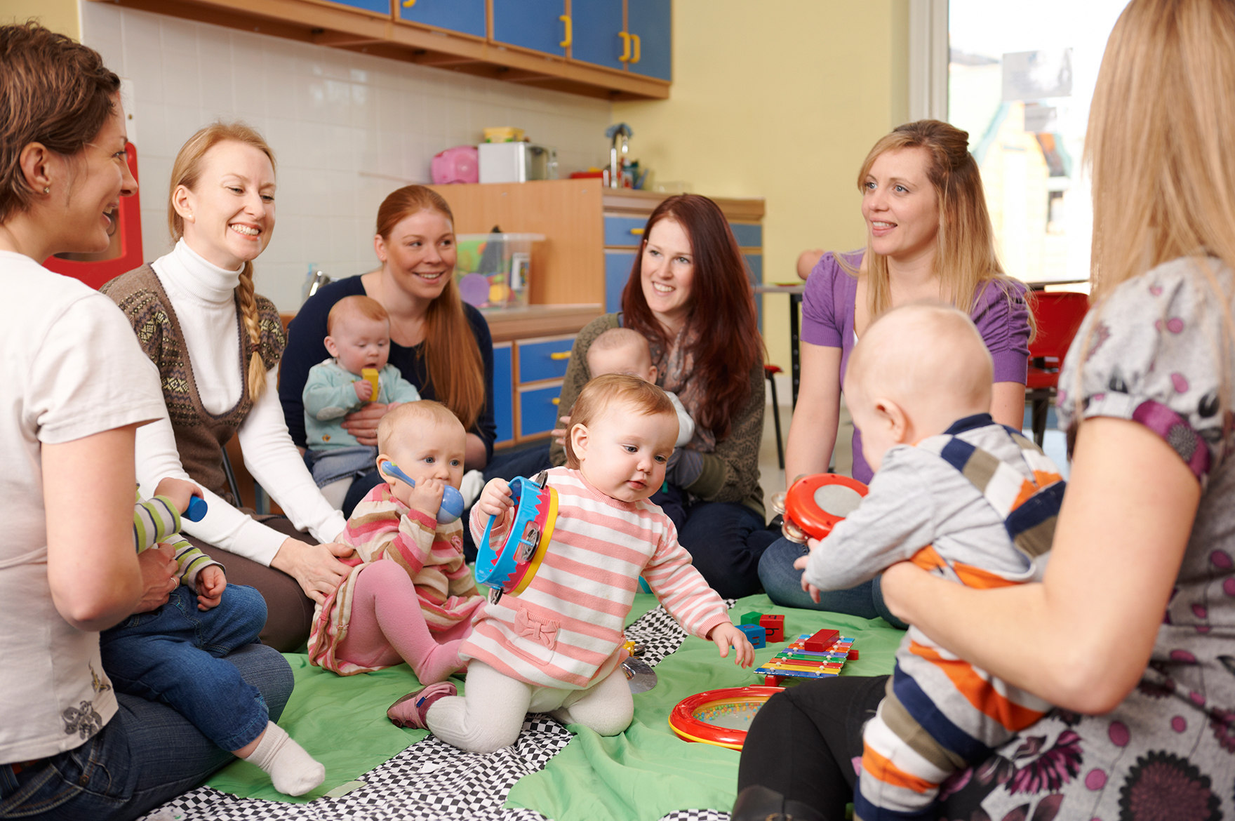 sechs junge Mütter sitzen mit ihren Babys im Kreis auf dem Boden und unterhalten sich