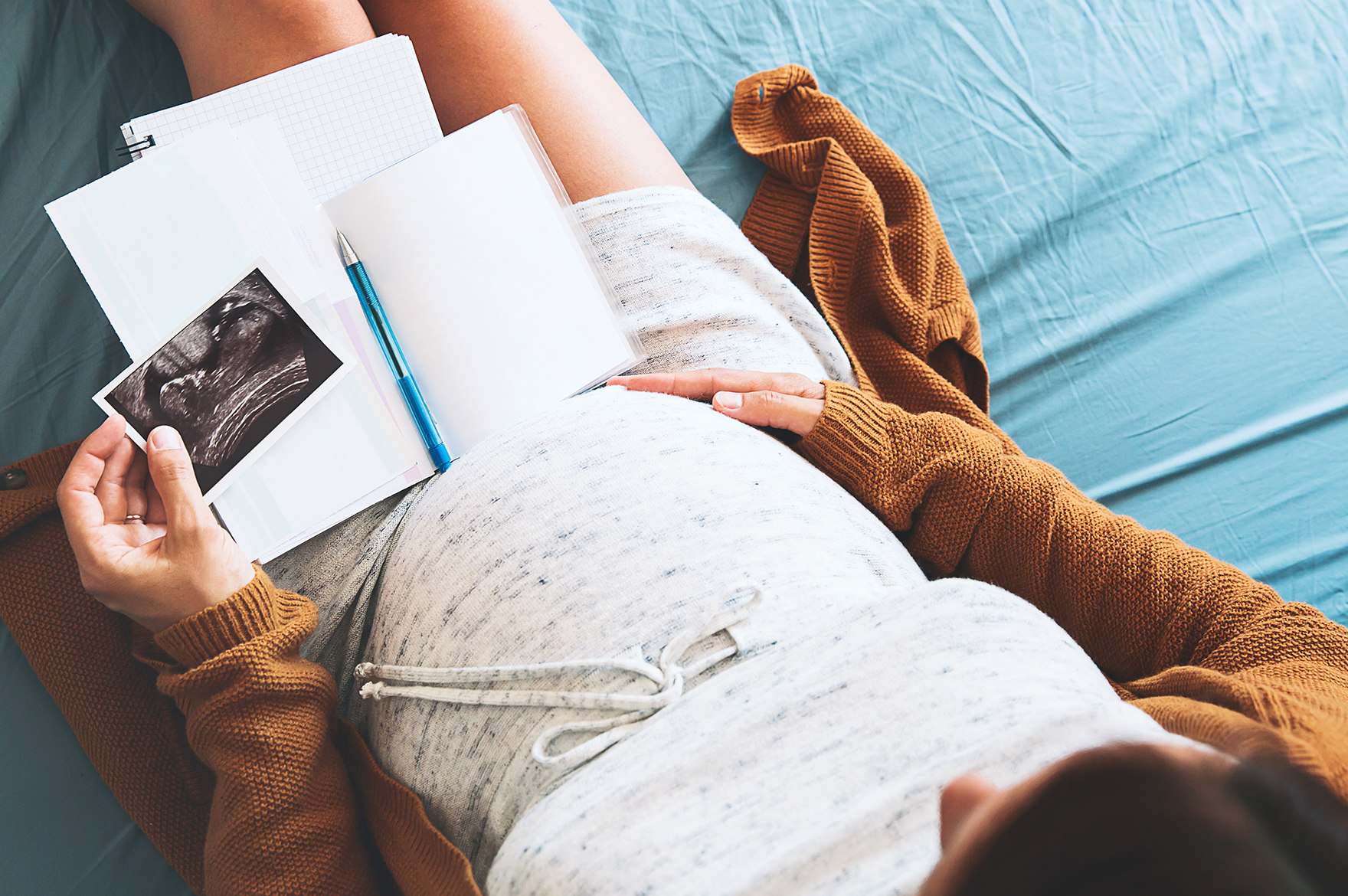 Eine Schwangere sitzt mit einem Notizbuch und Stift im Schoß auf dem Bett und hält in der linken Hand ein Ultraschallbild. Die rechte Hand liegt auf dem Bauch.