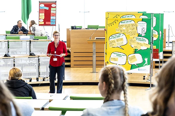 Die Kinder- und Jugendbeauftragte hält ein Mikrofon in der Hand und spricht im Landtag vor einer Schautafel mit bunten Zetteln