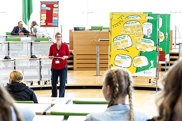 Die Kinder- und Jugendbeauftragte hält ein Mikrofon in der Hand und spricht im Landtag vor einer Schautafel mit bunten Zetteln