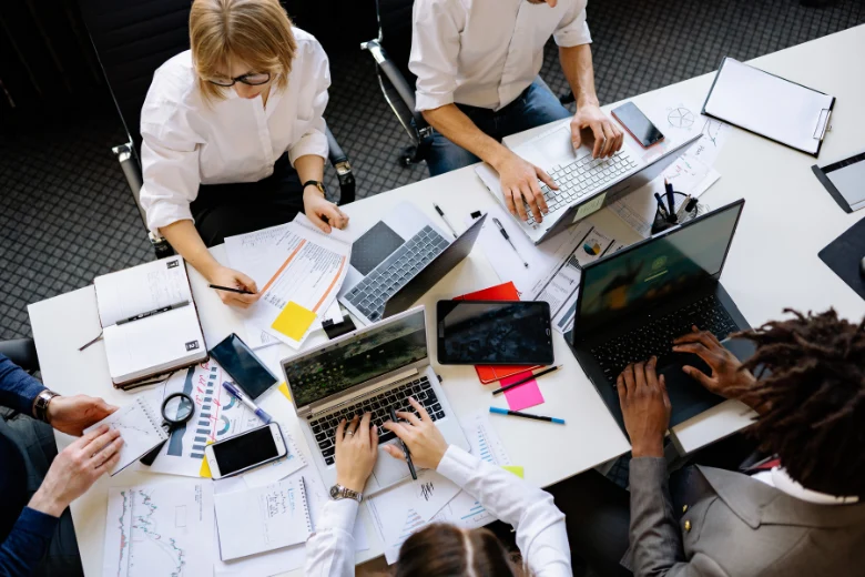 fünf arbeitende Jugendliche sitzen gemeinsam an einem Tisch und arbeiten an Computern, mit Handys und Notizen