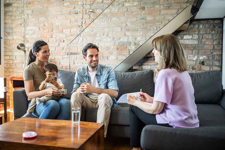 Beratungssituation: Junges Paar mit Kleinkind sitzt auf Sofa und spricht mit einer Therapeutin